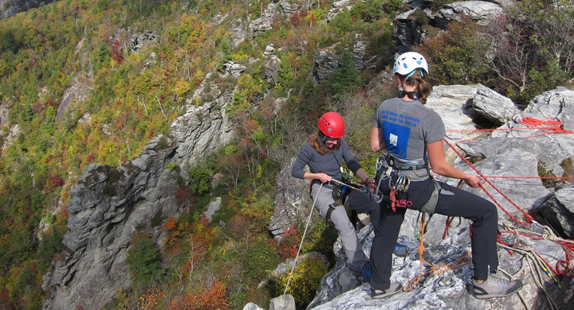 Two people wearing safety gear are secured by ropes near the edge of a cliff. One person appears to be an instructor, giving direction to the other person. Behind them is a rocky landscape covered in greenery. 